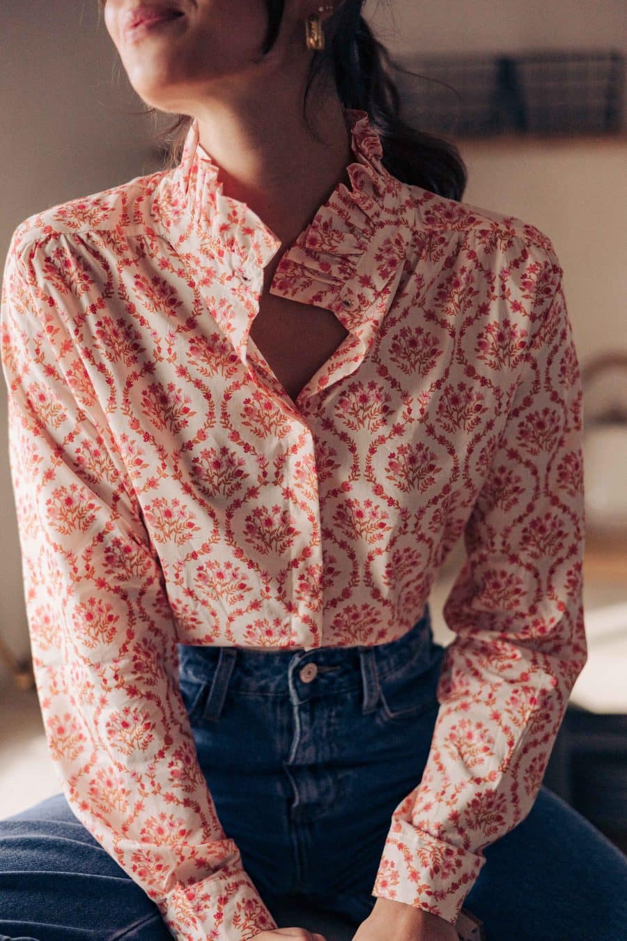 Chemise Maeline Sahoma écrue aux motifs floraux orangés, col montant à volants, manches longues, portée avec un jean bleu foncé