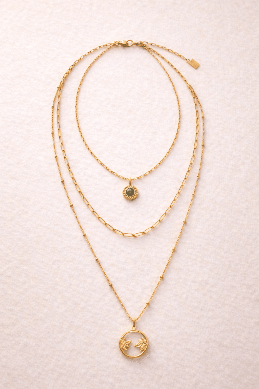 Collier Accord triple rang doré avec chaîne maillons à pendentif pierre verte, chaîne intermédiaire à maillons chunky ovales et longue chaîne perlée avec médaillon feuilles, sur fond beige texturé.