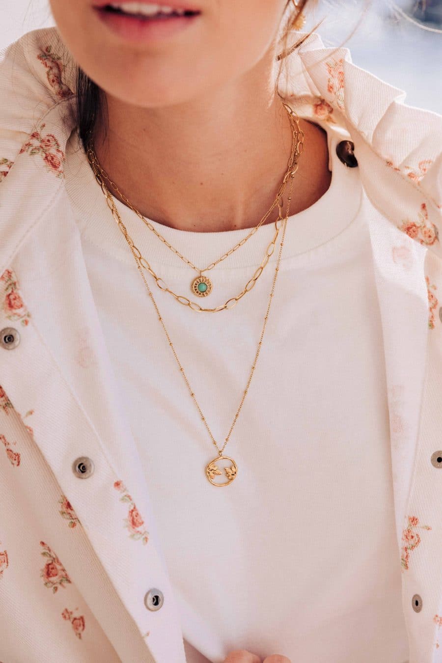 Collier Accord triple rang doré avec chaîne maillons à pendentif pierre verte, chaîne intermédiaire à maillons chunky ovales et longue chaîne perlée avec médaillon feuilles, sur fond beige texturé.